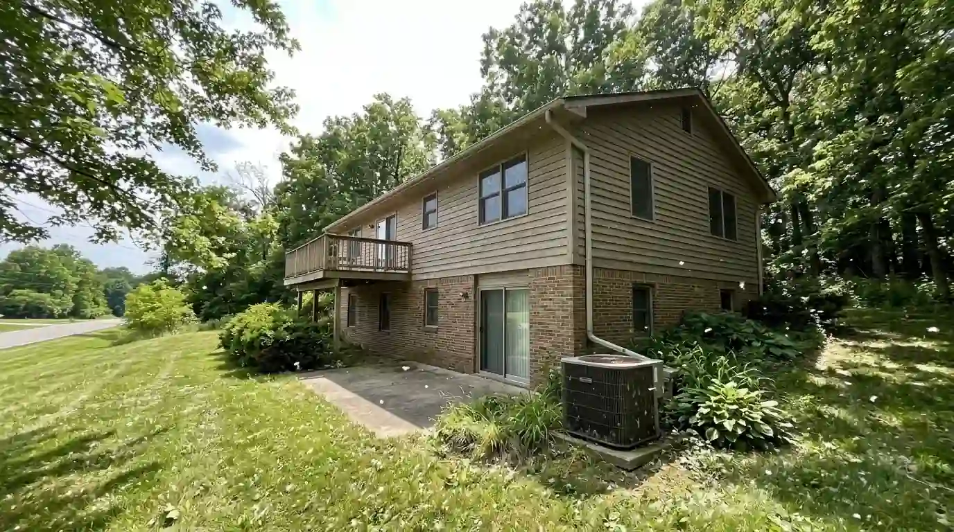 Two-story 1980s home on a heavily wooded lot along Covington Road in Fort Wayne, June afternoon, cottonwood fluff drifting over the outdoor AC unit by the patio.