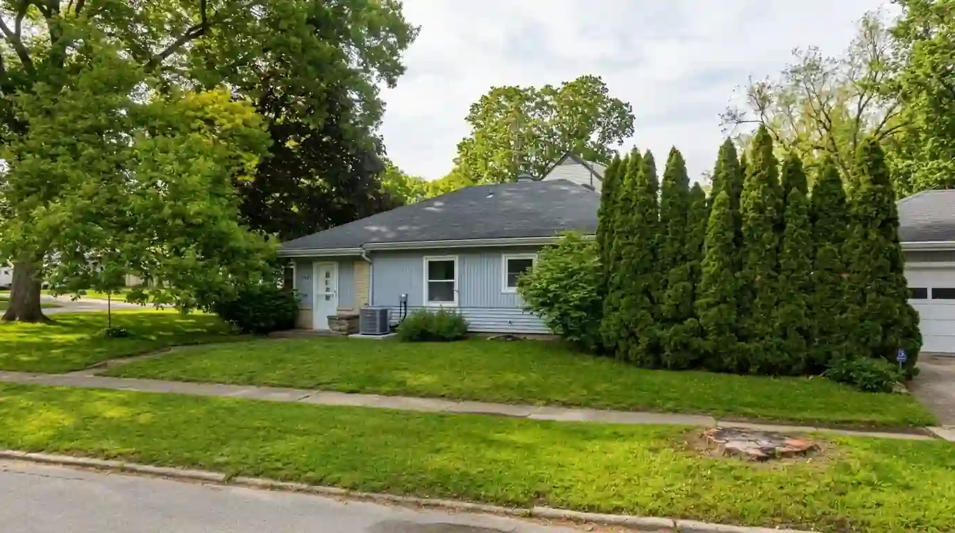 Small 1940s cottage-style home with a compact front yard in Indian Village, Fort Wayne, late spring morning, a new outdoor AC condenser beside the side door.