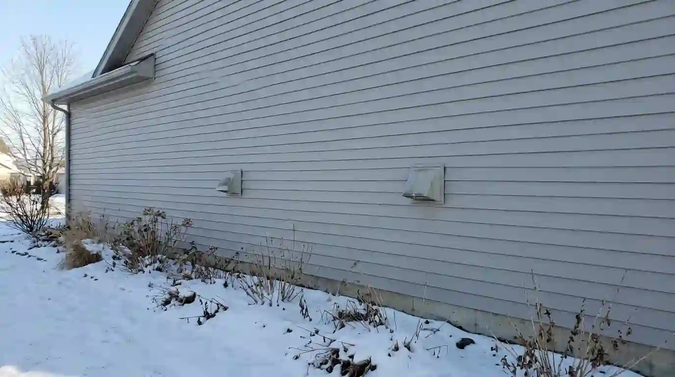 Two exterior ventilation hoods on the vinyl siding of a newer Huntertown, Indiana home, spaced well apart above a snowy flower bed, clear winter sky