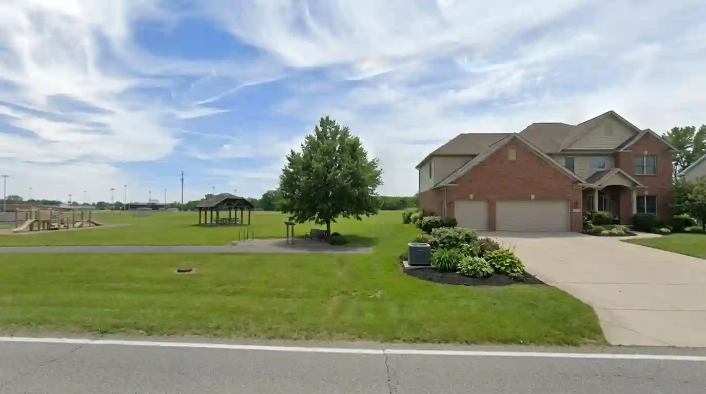 Large two-story suburban home with a three-car garage in Aboite, Fort Wayne, on a hot summer afternoon, outdoor AC condenser beside a mulched flower bed.