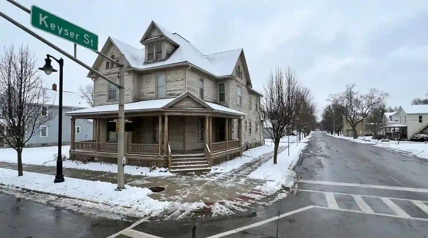 A tall early 1900s wood-frame house with a deep front porch on a quiet street in Garrett, Indiana, light snow on the roof, a winter afternoon.